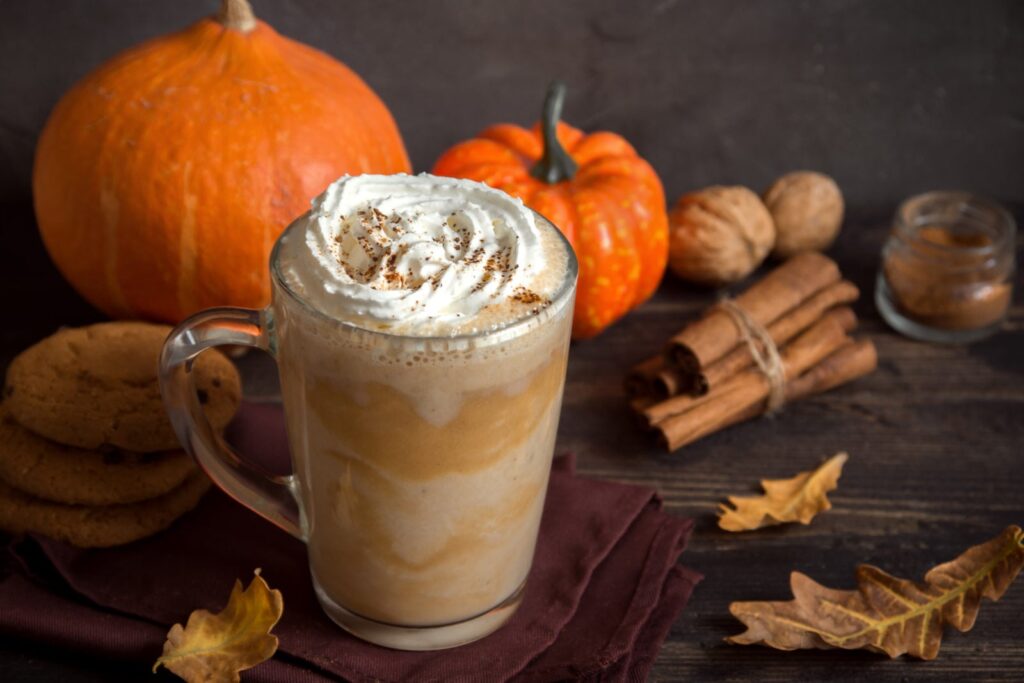 A cozy autumn setup featuring a pumpkin spice latte, pumpkins, and cinnamon sticks, highlighting early fall seasonal marketing to attract customers and build excitement.